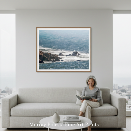A framed coastal landscape art print hangs on a white wall above a light gray sofa in a modern living room. A woman sits on the sofa reading a book. Seal Rocks San Francisco art by Murray Bolesta