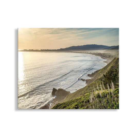 a serene coastal landscape with a rocky cliff overlooking a vast body of water, possibly a bay or ocean. The sky is filled with clouds, creating a dramatic backdrop for the scene. The sun is setting, casting a warm glow over the landscape and enhancing the colors of the scene. The image is a beautiful representation of nature's tranquility and the power of the ocean. Stinson Beach Sunset Art Print – Marin County California Coastal Decor