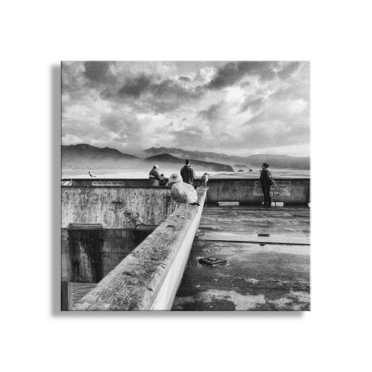 A black and white image depicting a group of people standing on a concrete structure, overlooking a body of water with mountains in the background. Pacifica Pier Black & White Art Print | California Coastal Wall Art