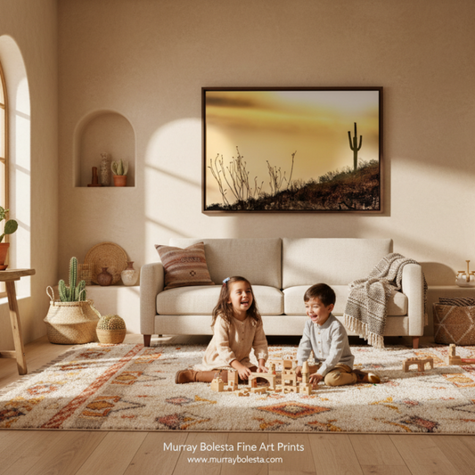 A framed landscape art print depicting a desert scene with a cactus under a warm golden sky, displayed on a beige wall in a beautifully styled living room with children playing on a patterned rug. By Murray Bolesta