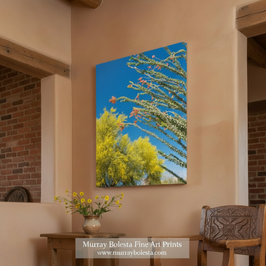 A vertical canvas wall art print depicting a desert landscape with green cactus branches and bright yellow-green trees under a vivid blue sky, displayed on a beige wall in a rustic room setting with a wooden table, chair, and brick archway. A watermark at the bottom reads 'Murray Bolesta Fine Art Prints'.