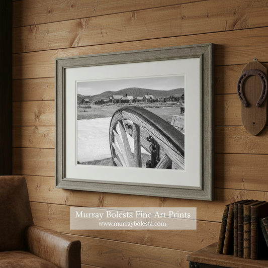Black and white photo of a wagon wheel with a ghost town in the. background. Bodie California by Murray Bolesta
