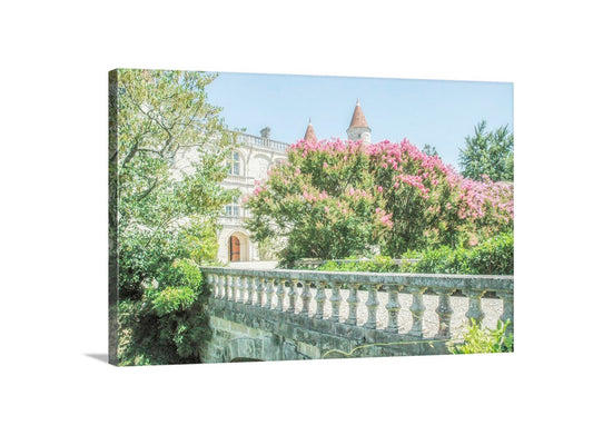 A stone bridge with a railing, surrounded by lush greenery and pink flowers, leading to a large, ornate building with a red roof and multiple towers. French Castle Photo Art Print | South of France Wall Decor