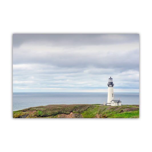 A white lighthouse stands on a grassy hill overlooking the ocean, with a cloudy sky in the background. Yaquina Head Lighthouse Art | Oregon Coast Minimalist Ocean Print
