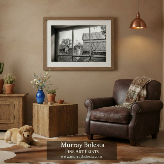 A framed black and white fine art print by Murray Bolesta hangs on a textured beige wall in a rustic living room setting with a brown leather armchair, wooden furniture, and a puppy on the floor.