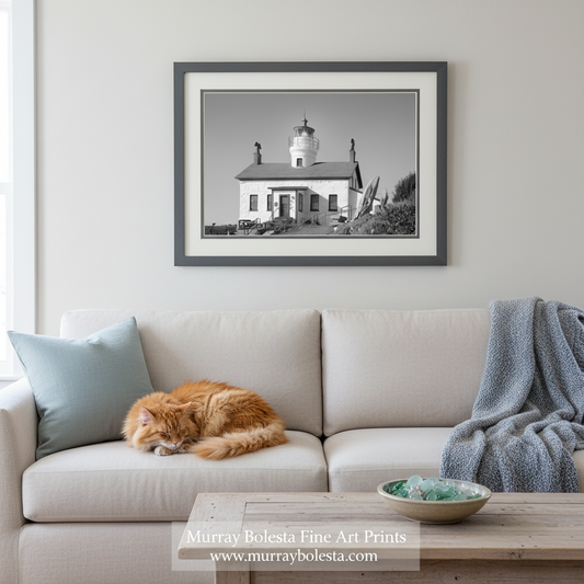 A framed black and white art print of a lighthouse hanging on a light gray wall above a beige sofa with a sleeping orange cat, a light blue pillow, and a gray throw blanket. A watermark at the bottom reads 'Murray Boleska Fine Art Prints'.