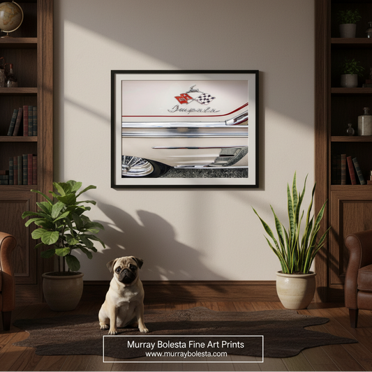 A framed art print featuring a vintage Chevy Impala car illustration, displayed on a beige wall in a styled room with a pug dog sitting on a rug below. The room includes wooden bookshelves, potted plants, and leather armchairs. By Murray Bolesta.