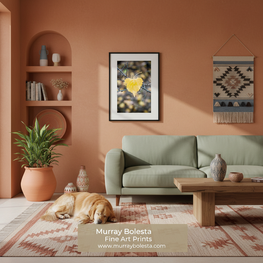 A framed art print featuring a yellow heart on a branch against a dark background, displayed on a terracotta wall in a styled living room with a green sofa, wooden coffee table, and a dog lying on a patterned rug. The text 'Murray Bolesta Fine Art Prints' is visible at the bottom.