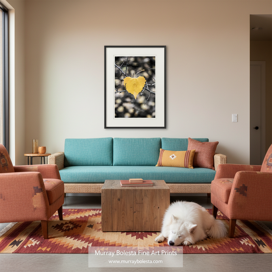 A framed art print featuring a yellow heart-shaped leaf on a branch against a dark background, displayed on a beige wall in a modern living room setting above a teal sofa with a white dog on a patterned rug.