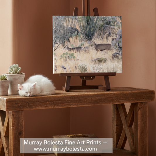 Southern Arizona landscape with mule deer, by Murray Bolesta. A square fine art print showing a desert landscape with deer in dry grass, displayed on a wooden easel atop a rustic wooden table. A white cat sleeps on the table beside small potted succulents.