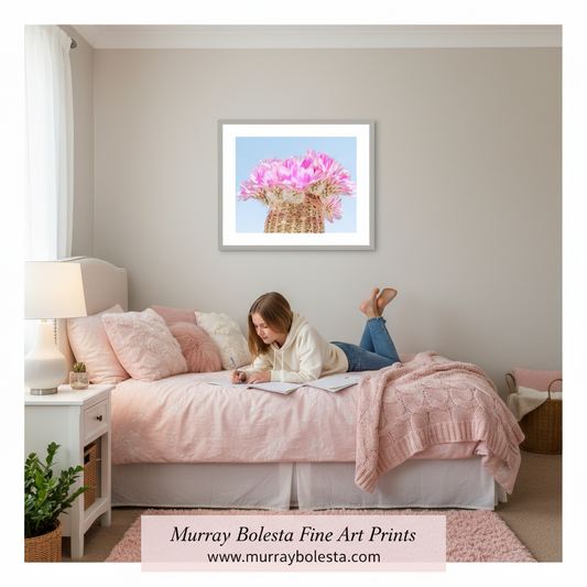 A framed art print featuring pink flowers in a basket against a light blue sky, displayed on a beige wall above a bed with pink bedding in a bedroom. A woman is lying on the bed writing in a notebook.