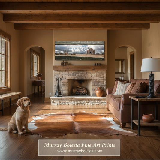 A framed landscape art print depicting a rustic farmhouse and barn under a cloudy sky, displayed above a stone fireplace in a warm, rustic living room with a dog sitting on the floor.