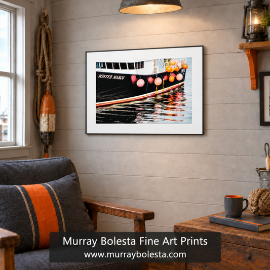 A framed art print of aboat hanging on a light gray wood-paneled wall in a cozy, rustic living room setting. The print features a black boat with orange and pink buoys and colorful reflections in the water. Boat art by Murray Bolesta