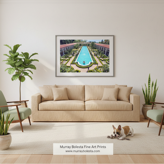 A framed art print depicting an aerial view of a luxurious swimming pool surrounded by manicured gardens and resort-style buildings, displayed on a light beige wall above a matching beige sofa in a modern living room setting. A small dog is lying on the rug in the foreground, and a brand watermark for 'Murray Boleta Fine Art Prints' is visible at the bottom.