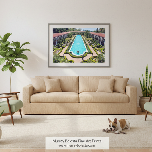 A framed art print depicting an aerial view of a luxurious swimming pool surrounded by manicured gardens and resort-style buildings, displayed on a light beige wall above a matching beige sofa in a modern living room setting. A small dog is lying on the rug in the foreground, and a brand watermark for 'Murray Boleta Fine Art Prints' is visible at the bottom.