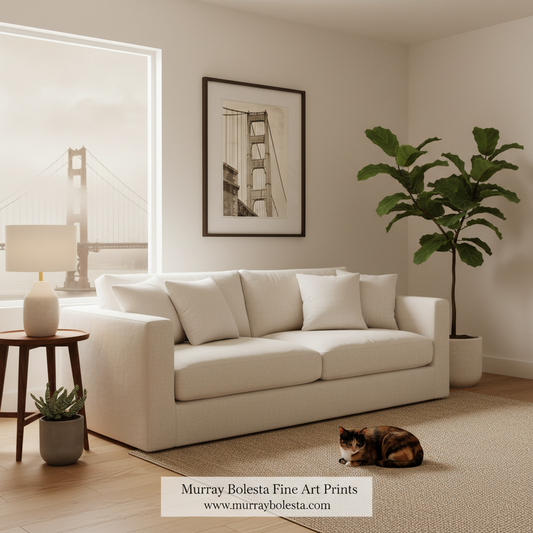 A bright, modern living room with a cream-colored sofa, a framed black-and-white art print of the Golden Gate Bridge on the wall, a large potted fiddle-leaf fig plant, and a small cat lying on the rug. A watermark at the bottom reads 'Murray Bolesta Fine Art Prints'.