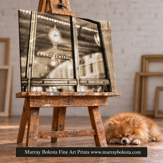 A framed art print featuring a sepia-toned Grand Central station scene, displayed on a rustic wooden easel in an art studio. A small fluffy dog is sleeping on the floor nearby, and a black banner at the bottom reads 'Murray Bolesta Fine Art Prints www.murraybolesta.com