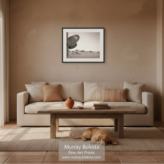 A framed black and white art print of a saguaro cactus and desert landscape hangs on a textured beige wall above a light beige sofa in a styled living room. A wooden coffee table, textured rug, and a sleeping dog are visible in the foreground.