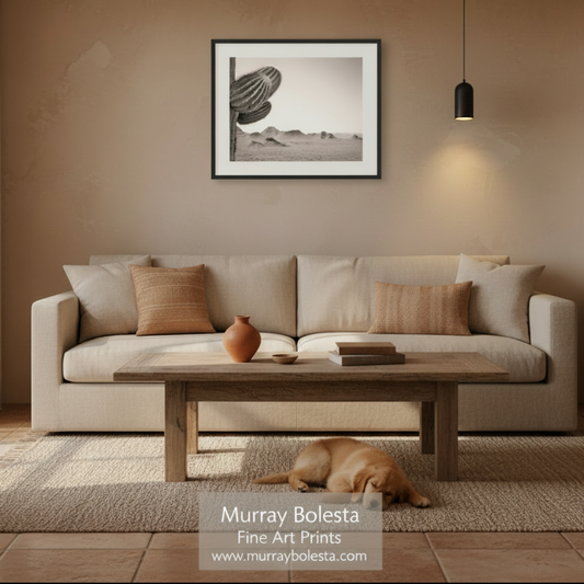 A framed black and white art print of a saguaro cactus and desert landscape hangs on a textured beige wall above a light beige sofa in a styled living room. A wooden coffee table, textured rug, and a sleeping dog are visible in the foreground.