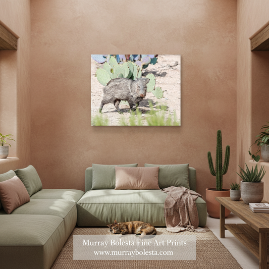 A square canvas art print depicting a wild boar walking among prickly pear cacti in a desert landscape, displayed on a warm terracotta wall in a modern living room setting above a sage green sectional sofa.