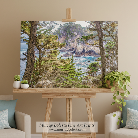 Point Lobos by Murray Bolesta. A large landscape art print depicting a coastal rocky shoreline with blue water, trees, and cliffs, displayed on a wooden easel in a styled living room setting with two beige armchairs and potted plants.