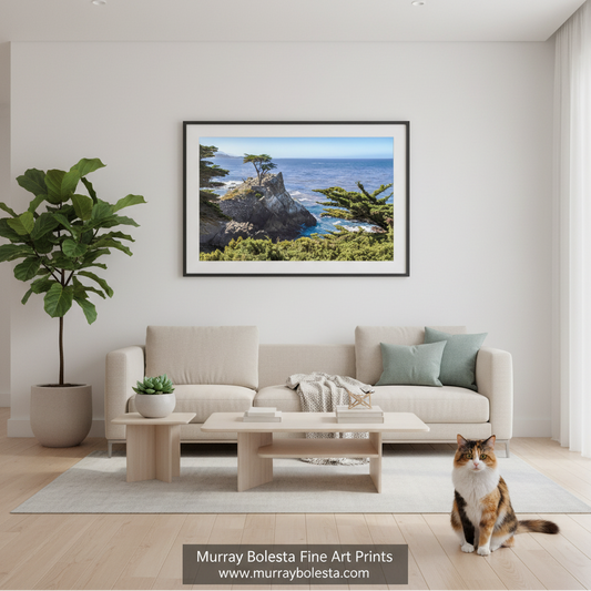Pebble Beach California by Murray Bolesta! A framed landscape art print of a coastal rocky cliff with a lone tree and ocean is displayed on a light gray wall above a beige sofa in a modern living room. A cat sits on the floor in the lower right, and a potted plant is on the left.