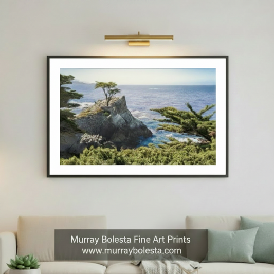 Pebble Beach California by Murray Bolesta! A framed landscape art print of a coastal rocky cliff with a lone tree and ocean is displayed on a light gray wall above a beige sofa in a modern living room. A cat sits on the floor in the lower right, and a potted plant is on the left.