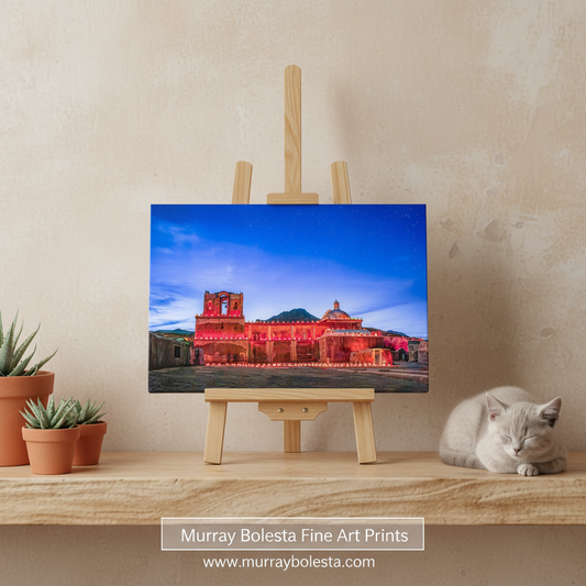 Tumacacori by Murray Bolesta. A framed-style art print showing a red-lit historic building under a blue sky, displayed on a small wooden easel on a tabletop. A gray cat rests at the right, and small potted succulents sit on the left against a textured beige wall.