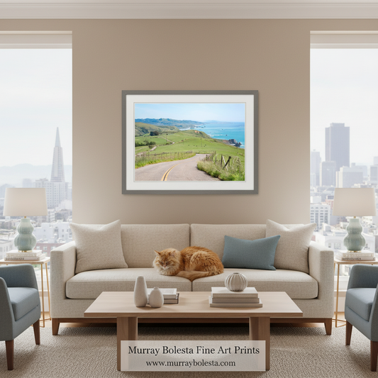A framed art print of a scenic coastal landscape with green hills, a winding road, and the ocean, displayed on a beige wall above a light-colored sofa in a modern living room setting. An orange cat is sleeping on the sofa.