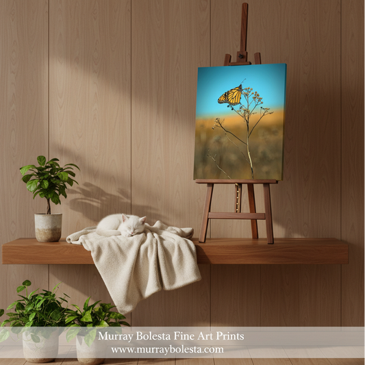 Monarch butterfly art! A framed-style canvas print showing a butterfly on a flowering branch against a blue and golden sky, displayed on a wooden easel in a styled interior with wood paneling, plants, and a draped cream blanket. Text at the bottom reads 'Murray Bolesta Fine Art Prints' and 'www.