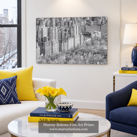 A black and white city skyline art print by Murray Bolesta is displayed on a white wall in a modern living room setting with yellow pillows, a marble coffee table, and a blue armchair. NYC winter art, upper west side.