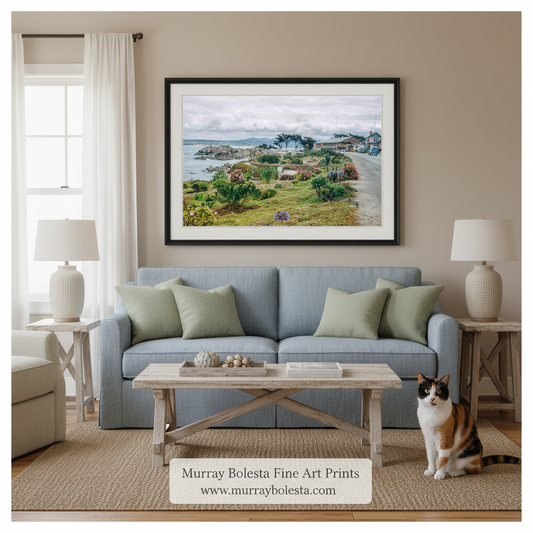A framed landscape art print by Murray Bolesta hangs on a beige wall above a light blue sofa in a styled living room. The print depicts a coastal village scene with green hills, water, and a cloudy sky.