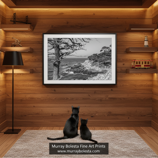 Seventeen Mile Drive, Pebble Beach California! A framed black and white coastal landscape art print hangs on a wood-paneled wall above two black cats sitting on a rug. The room has warm wood walls, shelves with small decor items, a floor lamp, and a visible watermark at the bottom reading Murray Bolesta Fine Art Prints.
