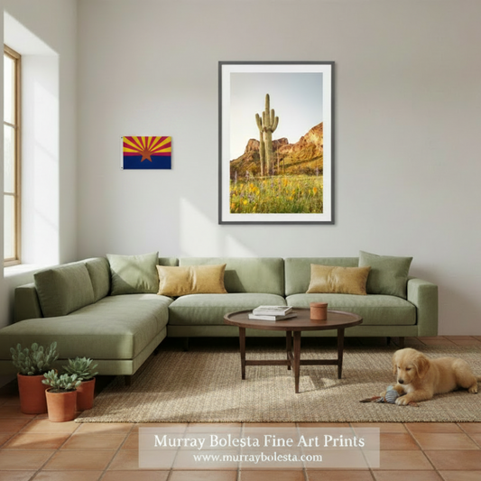 A framed art print of a desert landscape with a saguaro cactus and wildflowers, displayed on a light gray wall above a green sectional sofa in a styled living room. A golden retriever is lying on the rug on the floor.