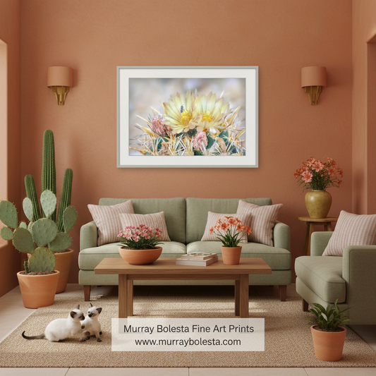 A framed floral art print featuring a yellow and pink cactus flower hangs on a terracotta wall above a green sofa in a styled living room. The print is the product being sold, but the image is a lifestyle mockup showing the artwork in a real interior setting with plants, furniture, and a cat.