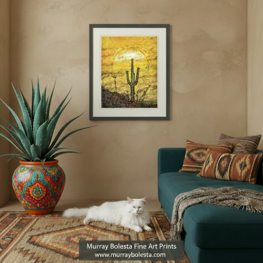A framed art print depicting a desert sunset with saguaro cacti, displayed on a beige wall in a styled living room with a teal sofa, patterned pillows, potted cacti, and a white cat on a rug. A watermark at the bottom reads 'Murray Boleta Fine Art Prints'.