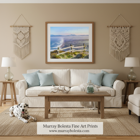 A framed landscape art print by Murray Bolesta hangs on a beige wall above a white sofa in a styled living room. The print depicts a coastal scene with blue water, distant mountains, and a wooden fence.