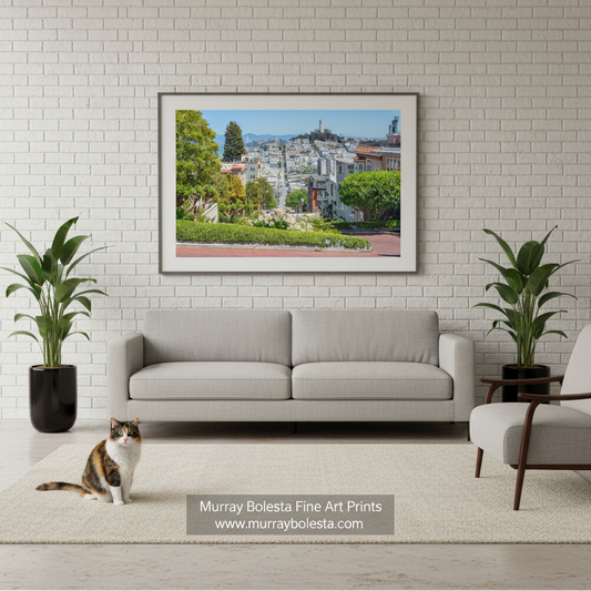 San Francisco Lombard Street by Murray Bolesta! A framed art print of a scenic hillside town with green trees and blue sky, displayed on a white brick wall above a light gray sofa in a modern living room. A cat sits on the rug in the foreground, and a plant is on each side.