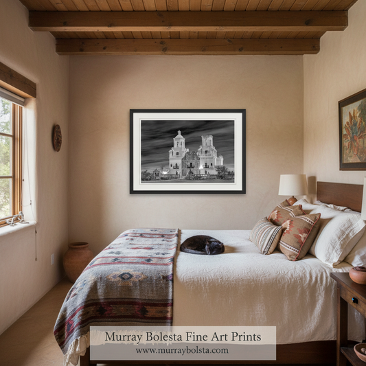 A black and white fine art photograph of a historic Spanish colonial church framed in black with a white mat, displayed on a beige wall above a bed in a rustic bedroom setting. A watermark for 'Murray Bolesta Fine Art Prints' is visible at the bottom.