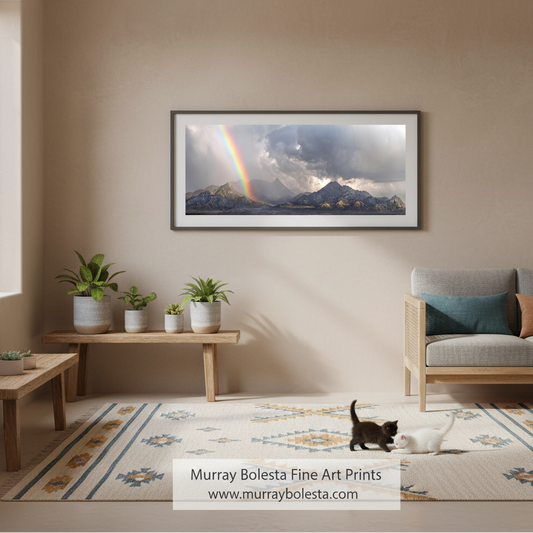A framed landscape art print of a mountain scene with a rainbow hangs on a beige wall in a styled living room with a sofa, bench, plants, and two cats on a rug. Text at the bottom reads 'Murray Bolesta Fine Art Prints' with the website.