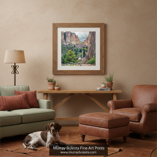 A framed art print depicting a scenic canyon landscape with green trees and a bridge, displayed on a beige wall in a styled living room with a sofa, armchair, ottoman, and a dog lying on the floor. A watermark at the bottom reads 'Murray Bolesta Fine Art Prints'.