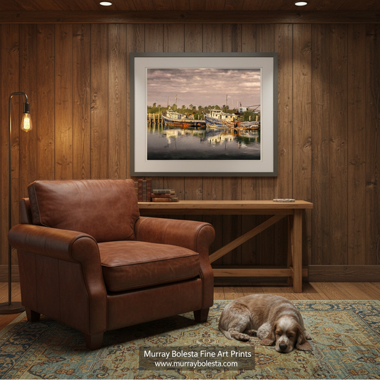 South Padre Island Texas. Murray Bolesta. A framed art print depicting a calm harbor scene with boats and reflections, displayed on a wood-paneled wall above a brown leather armchair and a sleeping dog in a rustic room.