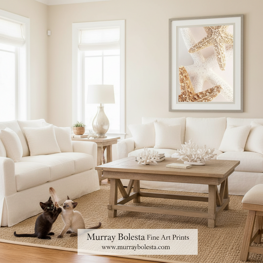 A styled living room with white sofas, a wooden coffee table, and a large framed art print on the wall featuring a starfish design. Two cats are on the floor near the table, and a text overlay at the bottom reads 'Murray Bolesta Fine Art Prints www.
