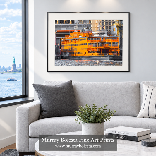 A framed art print of an orange ferry in a harbor is displayed on a light gray wall above a modern light gray sofa in a living room setting. The print is shown in a styled interior mockup with a window view of the Statue of Liberty. Ferry art by Murray Bolesta