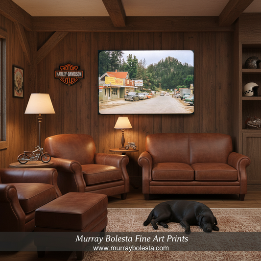 A framed art print depicting a vintage road scene in a small town hangs on a dark wood-paneled wall in a rustic living room. The room features brown leather sofas, armchairs, a matching ottoman, a black dog lying on the rug, and a Harley-Davidson logo on the wall.