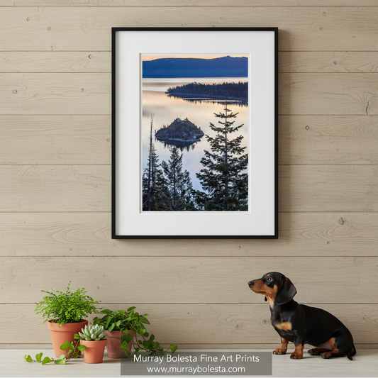 a serene lake surrounded by a forested island, with a mountain range in the background. The sky is painted in shades of orange, creating a beautiful contrast with the calm waters of the lake. Fine Art Tahoe Sunrise Print | Emerald Bay Tree Silhouette Vertical Art by Murray Bolesta!