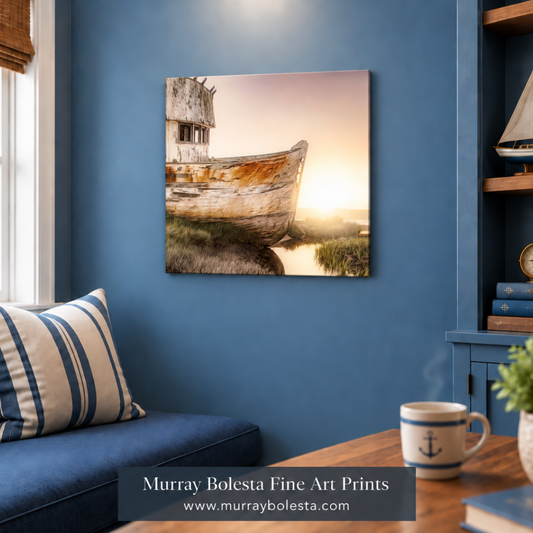 A square canvas wall art print depicting an old weathered boat on a marshy shore at sunset, displayed on a blue wall in a styled nautical-themed room with a sofa, striped pillow, wooden shelf, and a mug in the foreground. Tomales Bay art by Murray Bolesta