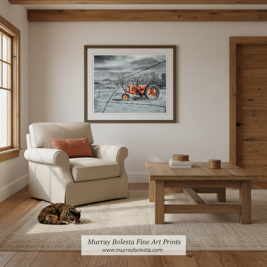 A framed art print depicting an orange tractor in a snowy field, displayed on a beige wall in a styled living room with a cream armchair, wooden coffee table, and a sleeping cat on the rug.
