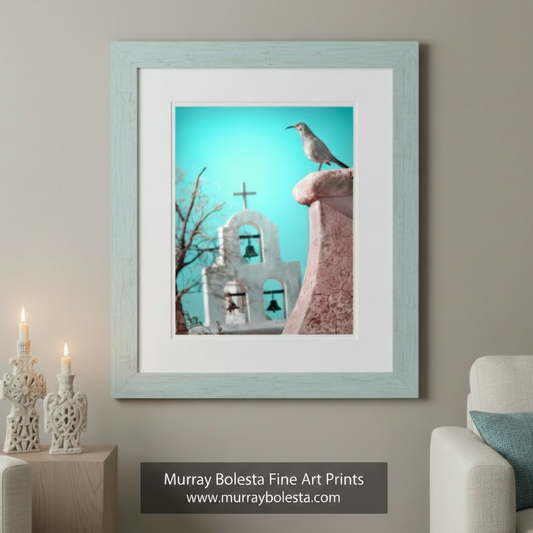 A bird perches on a pink stone structure, with a white church featuring a cross and bell in the background. Surreal Bird Art | San Xavier Mission Chapel Tucson Arizona Print by Murray Bolesta