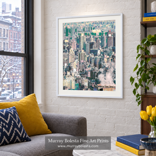 A framed art print of a dense city skyline hangs on a white brick wall in a modern living room setting with a gray sofa, yellow pillows, and a potted plant. NYC winter art by Murray Bolesta.